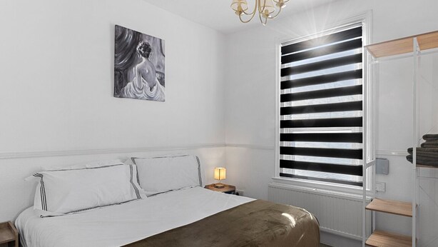 A bedroom with a bed, a nightstand, a lamp, a window with blinds, a towel rack, and a painting on the wall. - Central Exeter Apartment Kimberley Road (Exeter)