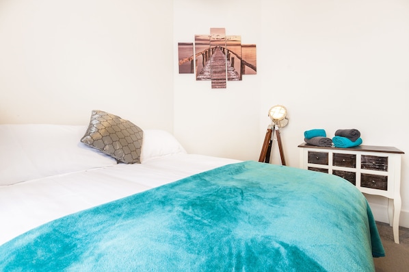 A neatly made bed with a teal blanket, a patterned pillow, and a wall-mounted picture of a pier.