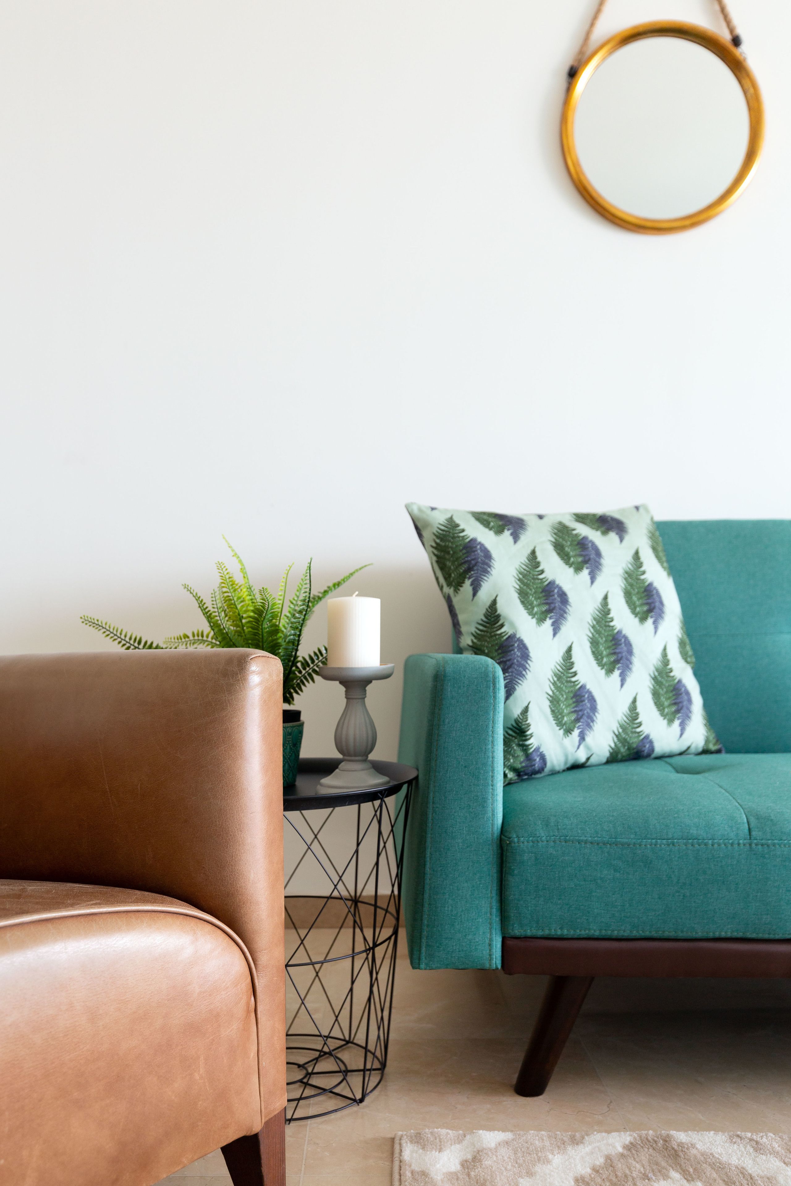 A teal sofa with a patterned pillow, a brown leather chair, a small side table with a candle, and a potted plant.