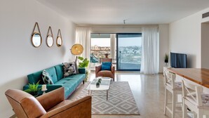 Ein modernes Wohnzimmer mit einem petrolfarbenen Sofa, einem braunen Ledersessel, einem Glastisch und einem Holzes Esstisch. An der Wand hängen drei runde Spiegel, eine stehende Stehlampe und ein Blick auf die Stadt durch große Fenster.