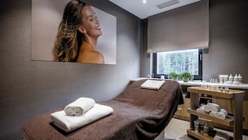 Un moderno spa con una camilla de masaje, toallas, un lavamanos y un ventanal con vistas a árboles.