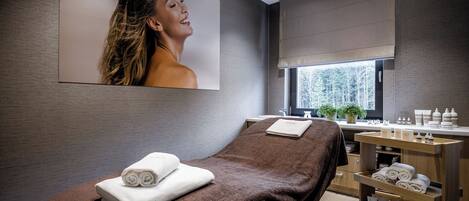 A modern spa room with a massage table, towels, a sink, and a window with a view of trees.