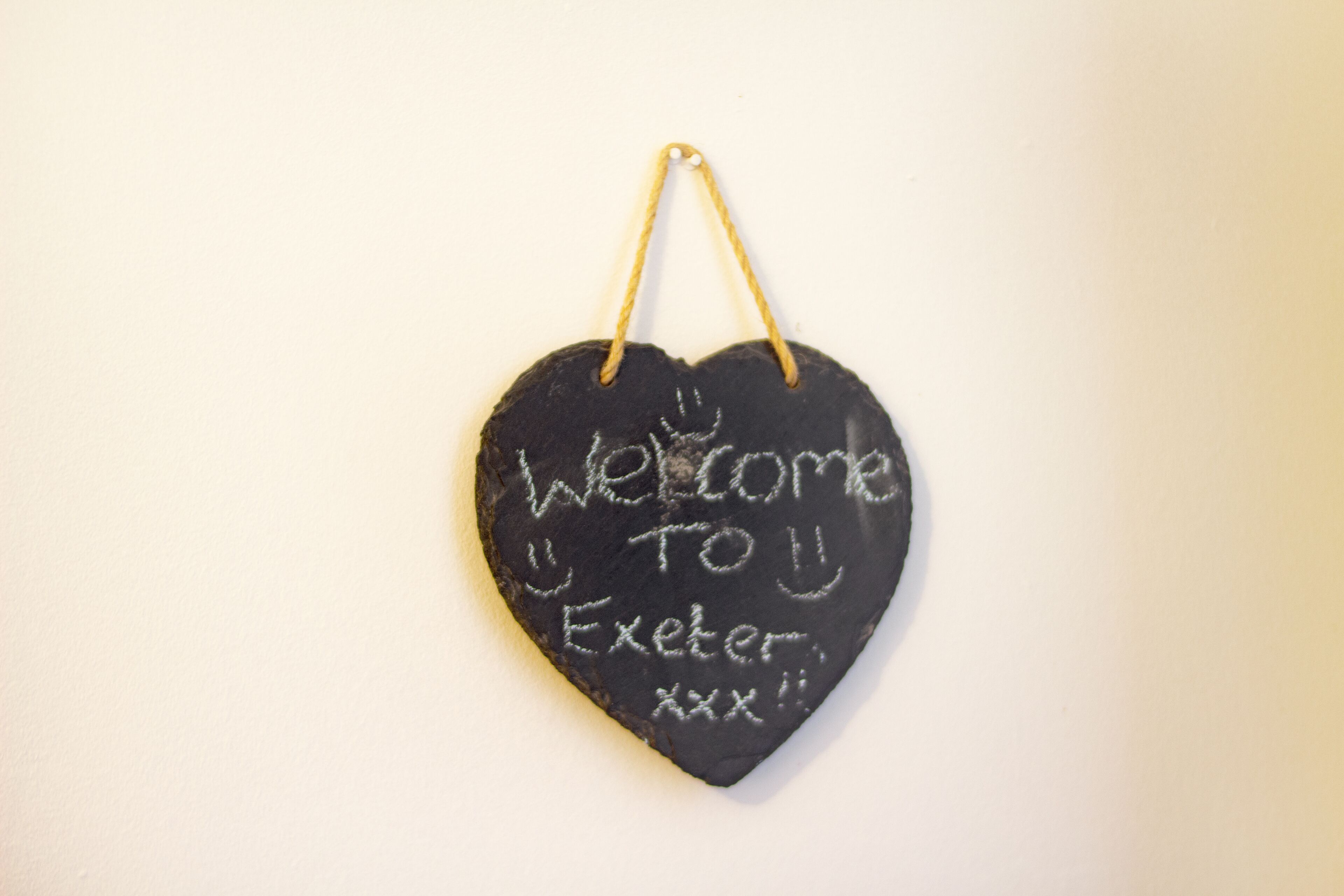 Um quadro-negro em formato de coração com a frase 'Welcome to Exeter' escrita, pendurado numa parede.