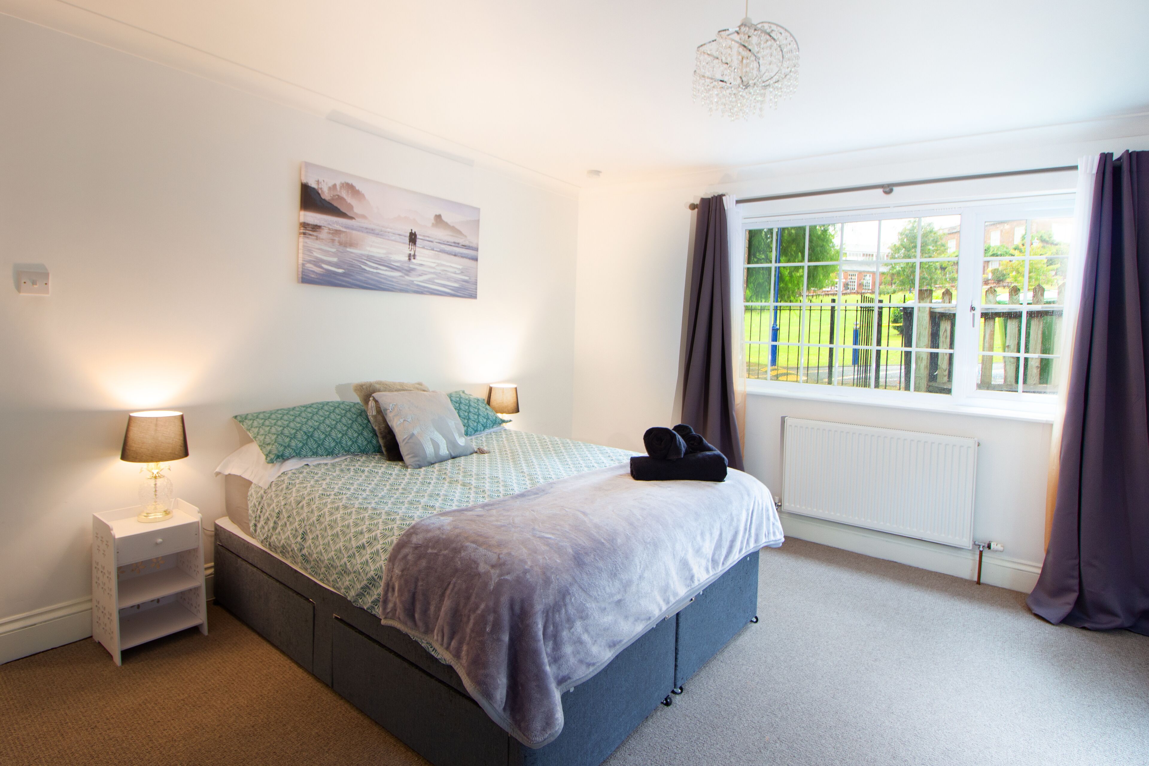 A bedroom with a bed, a nightstand, a window with curtains, and a radiator.