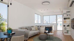 A modern living room with a grey sofa, a round dining table, and a small round table. There is a large window providing a view of the city. - Magic Penthouse TLV Harbor (Tel Aviv)