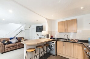 A compact kitchen with a washing machine, wooden cabinets, a bar stool, and a sofa with cushions. - Modern Exeter City Cottage Howell Road (Exeter)
