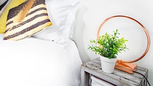 Un lit bien fait, avec du linge de lit blanc et des coussins décoratifs, une petite table de chevet en bois sur laquelle se trouve une plante en pot et un miroir rond, et un mur blanc en arrière-plan.