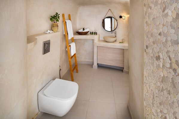A bathroom with a white toilet, a wooden ladder, a shelf with toiletries, a round mirror, and a pebble-patterned wall. - Aegean Mist (Santorini)