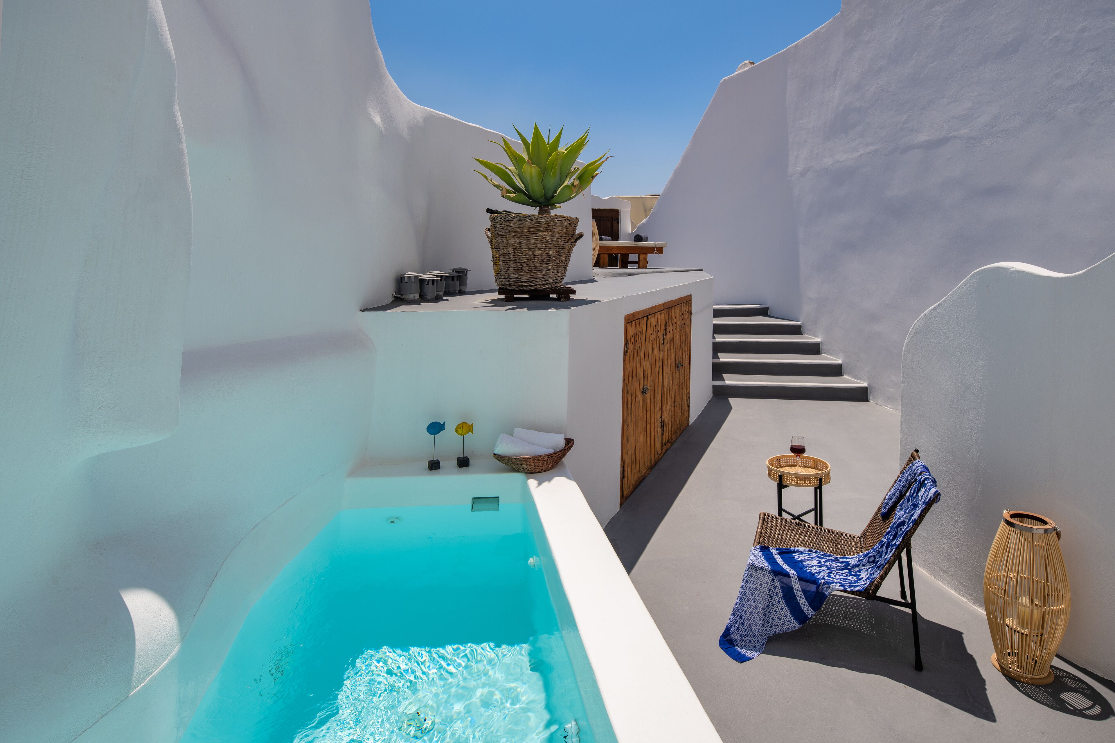 Un patio estrecho con paredes blancas, una piscina infinita azul, una silla de mimbre, una mesa pequeña con un vaso y una planta grande en maceta.