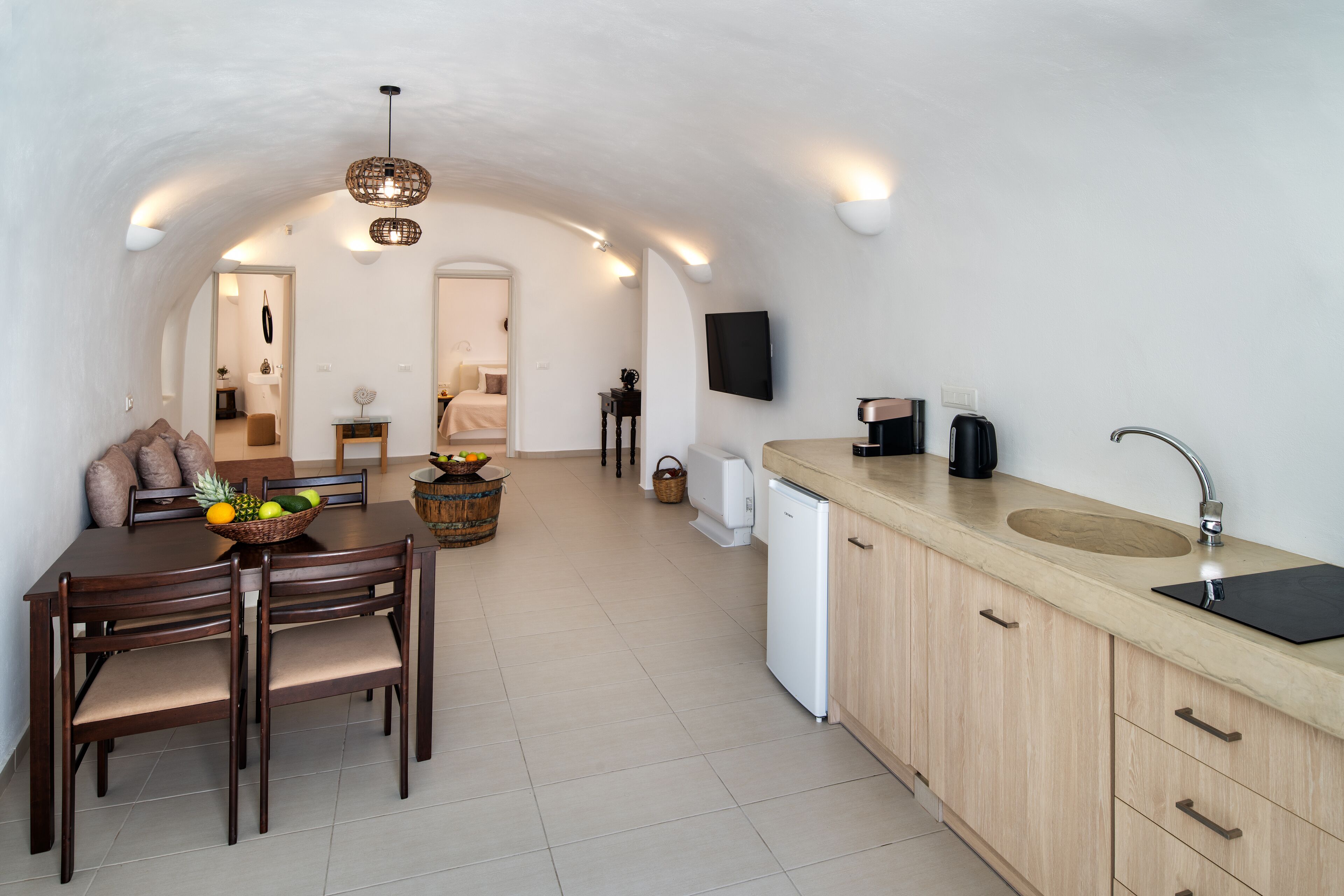 A modern kitchen with a wooden dining table, chairs, and a fruit bowl. There is a white refrigerator, a sink with a faucet, and a television mounted on the wall.