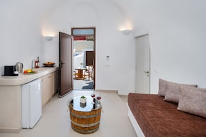A compact living space with a kitchenette, a round wooden table with a plant and wine glasses, a sofa with cushions, and a view into an adjacent room with a dining table and chairs.