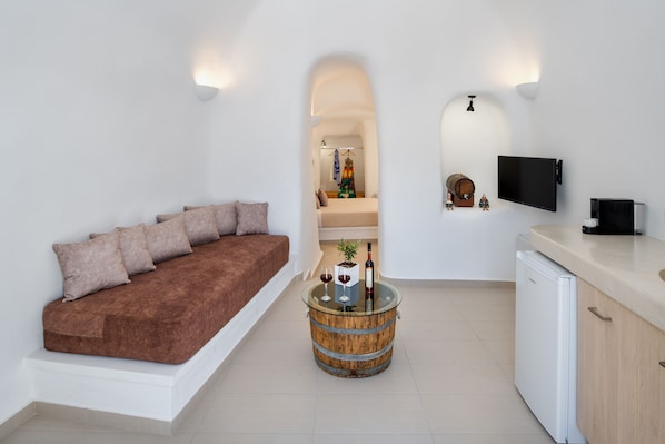 A modern living room with a sofa, a glass-top coffee table, a television, and a kitchen area. - Aegean Mist (Santorini)