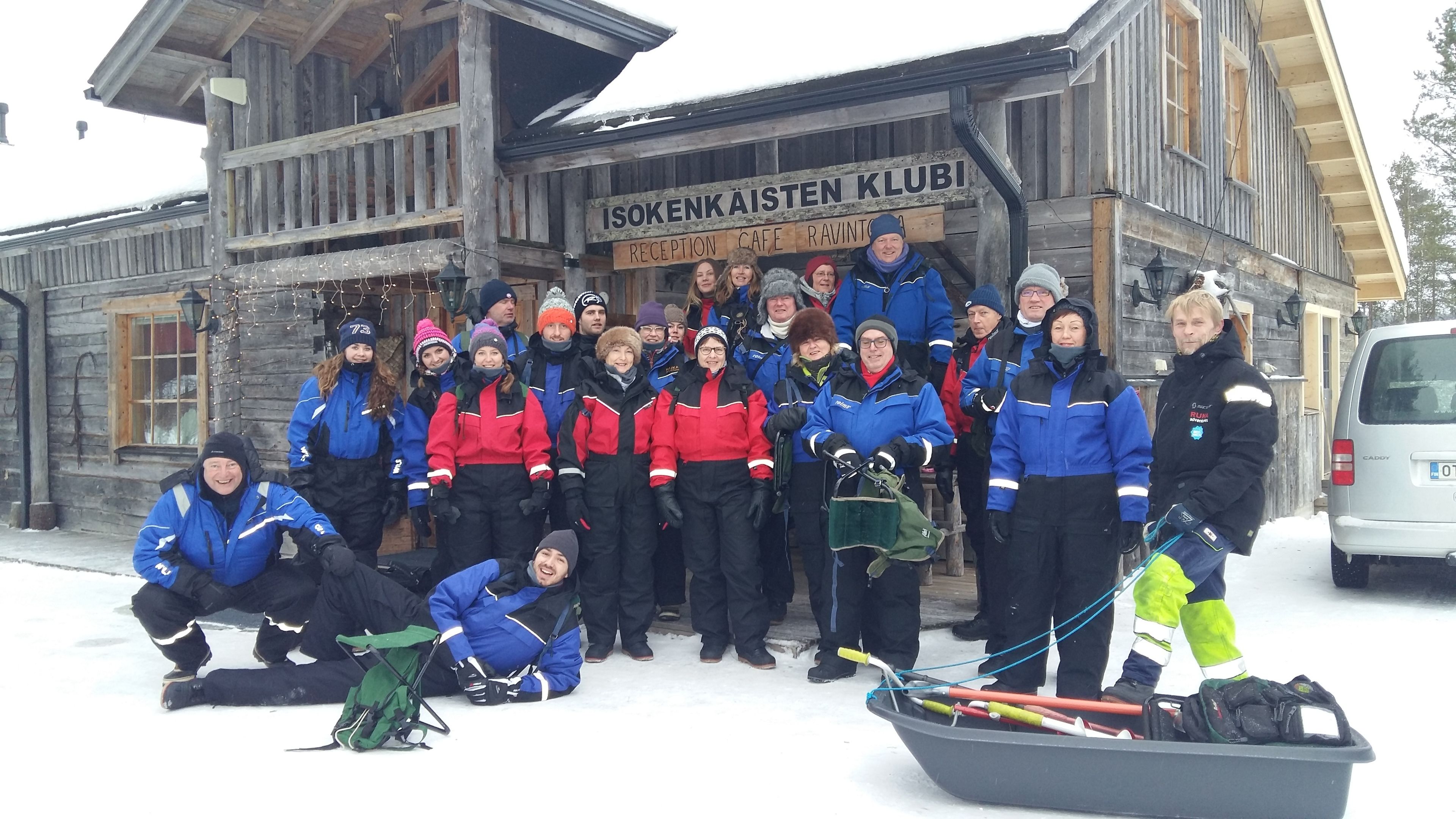 Een groep mensen in winterkleding poseert voor een houten gebouw met een bord waarop staat 