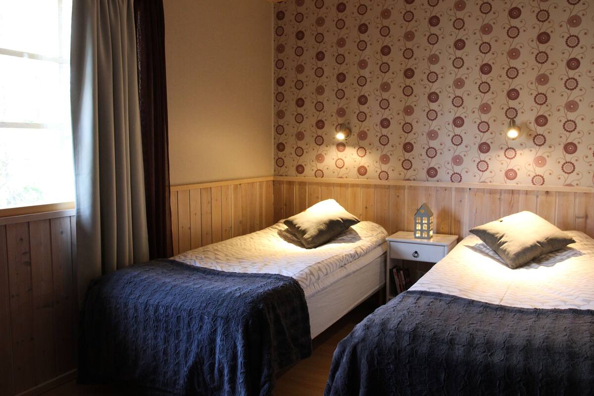 A room with two single beds, wooden walls, a patterned wallpaper, and a nightstand with a small house-shaped lamp.