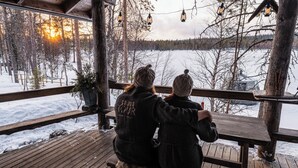 Two people sitting on a wooden bench overlooking a snowy landscape with a wooden deck and hanging lanterns. - Wilderness chalet Kuusamo (Kuusamo)