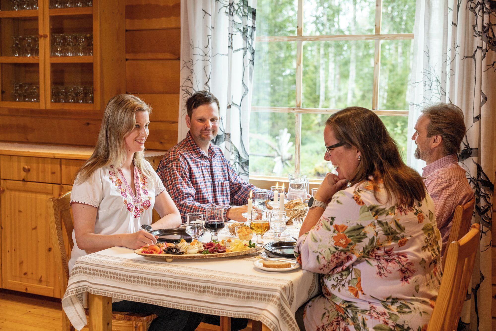 A wooden dining table with a white tablecloth, surrounded by wooden cabinets and a window with a view of trees.