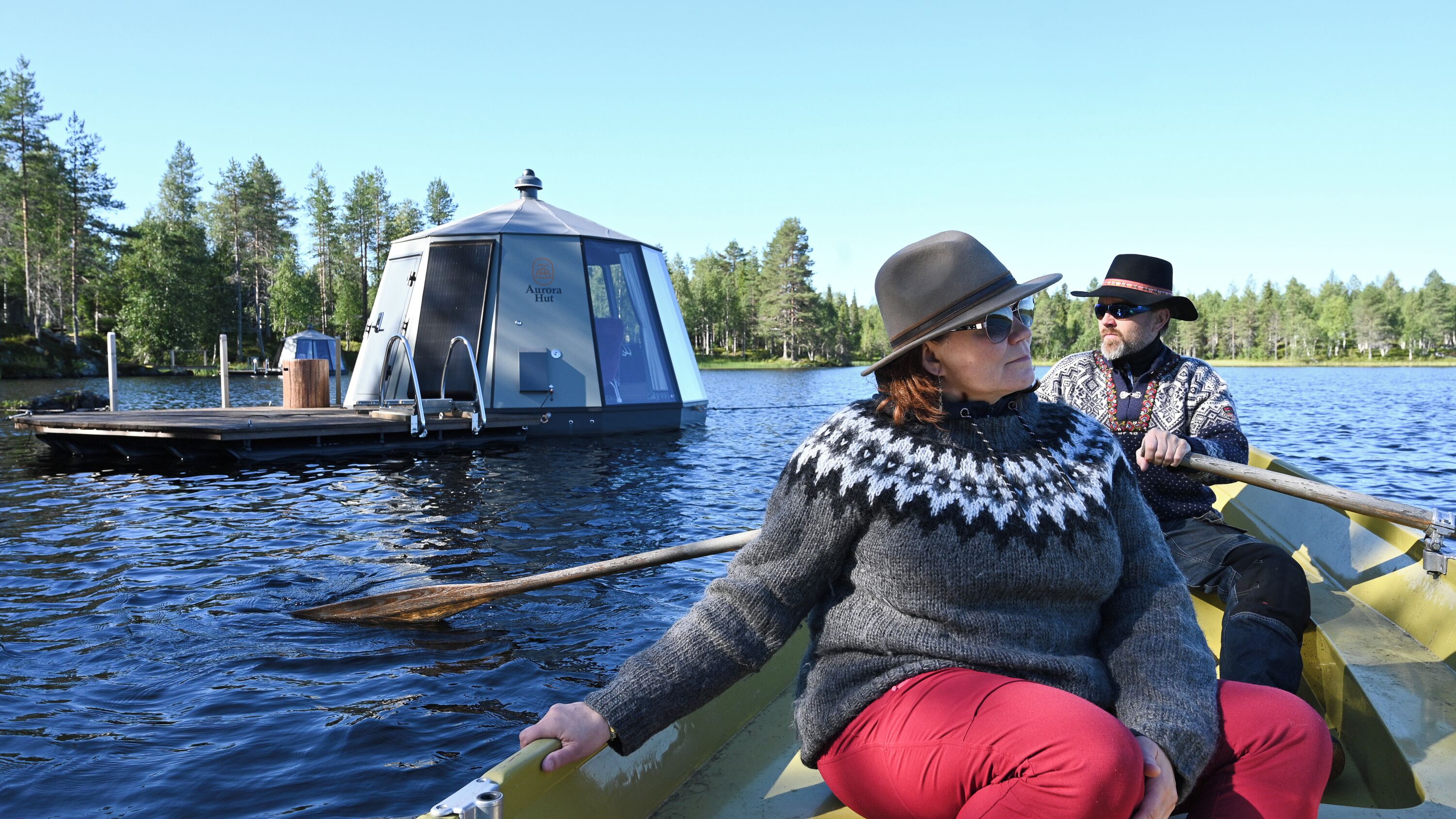 Un couple est assis dans un bateau à rames sur un lac, avec une cabane Aurora amarrée en arrière-plan.