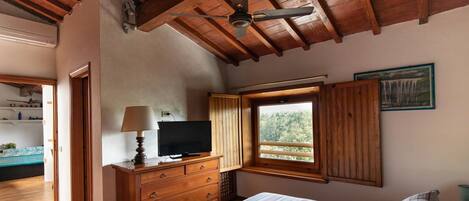 Un dormitorio con techo de madera, una cama con ropa de cama a rayas, un televisor sobre un aparador de madera y una ventana con vista a árboles.