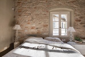 Une chambre avec un mur en pierre, une grande fenêtre, un lit recouvert de draps blancs et deux lampes de chevet.
