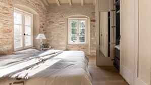 Un dormitorio con pared de piedra, una cama con cubrecama blanco, una ventana con vista a árboles, y un armario con espejo.