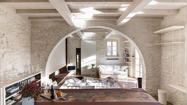 Amplia habitación con techo alto, paredes de piedra y un gran arco de entrada. Hay una zona de estar acogedora con un sofá y una mesa de comedor con copas de vino, pan y una botella.