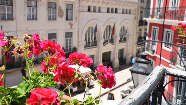 Uma varanda com flores vermelhas, um edifício branco com janelas em arco e um edifício vermelho com varandas.