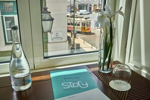 Un folleto del directorio del hotel, una botella de agua y un jarrón con flores blancas sobre una mesa junto a una ventana.