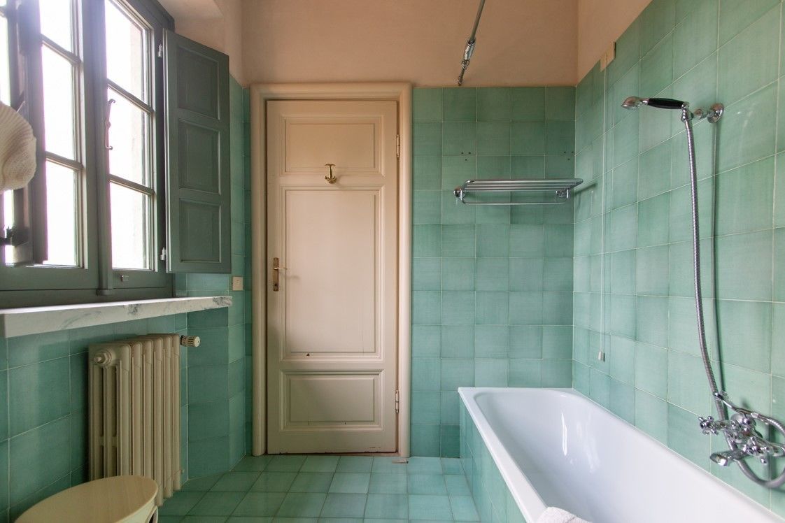 A bathroom with a bathtub, a radiator, a door, and a window with a towel hanging on the sill.