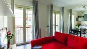 Una sala moderna con un sofá rojo, ventanas amplias con cortinas, un comedor con mesa y sillas, y una cocina al fondo.
