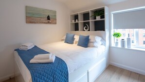 A neatly made bed with a blue quilt, white pillows, and a blue blanket. There is a painting on the wall, a shelf with decorative items, and a window with a plant. - Alison Court (Bristol)
