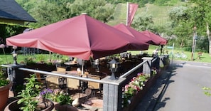 An outdoor seating area with tables and chairs under red umbrellas, surrounded by potted plants and flowers. - Moselperle (Traben-Trarbach)