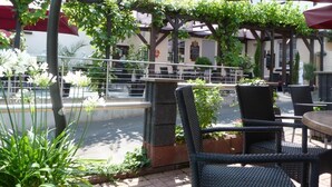 An outdoor seating area with wicker chairs and red umbrellas, surrounded by greenery and a building in the background. - Moselperle (Traben-Trarbach)