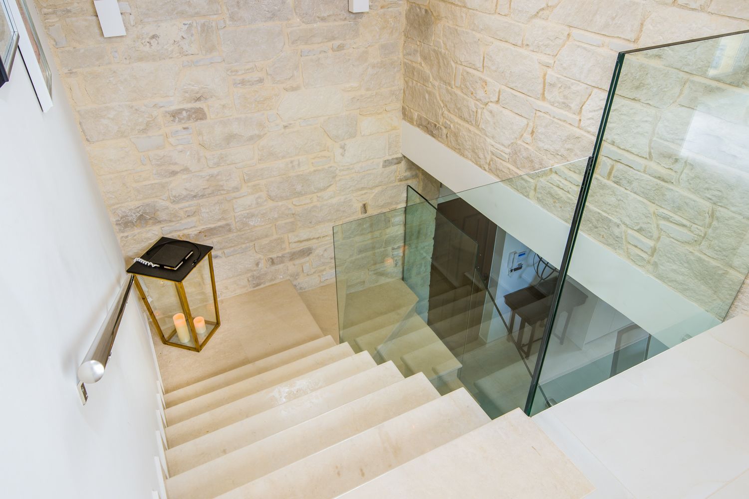 A modern staircase with glass railings, stone walls, and a small table with a candle.