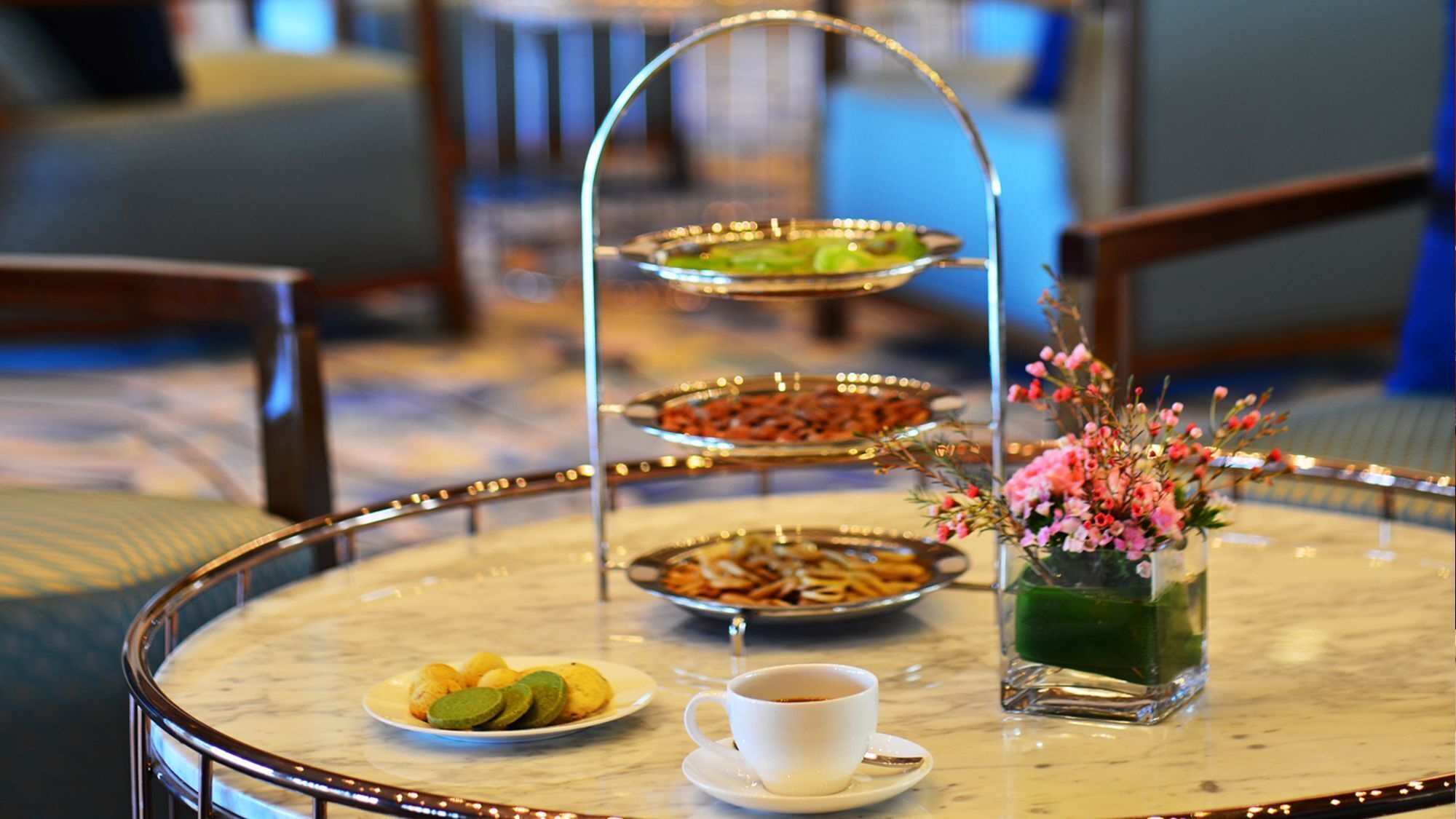 A three-tiered dessert stand with various sweets, a cup of coffee, and a floral arrangement on a round table.