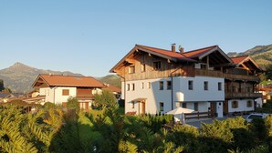 Une maison en bois à deux étages, dotée d’un balcon, entourée d’une végétation luxuriante et avec des montagnes en arrière-plan.