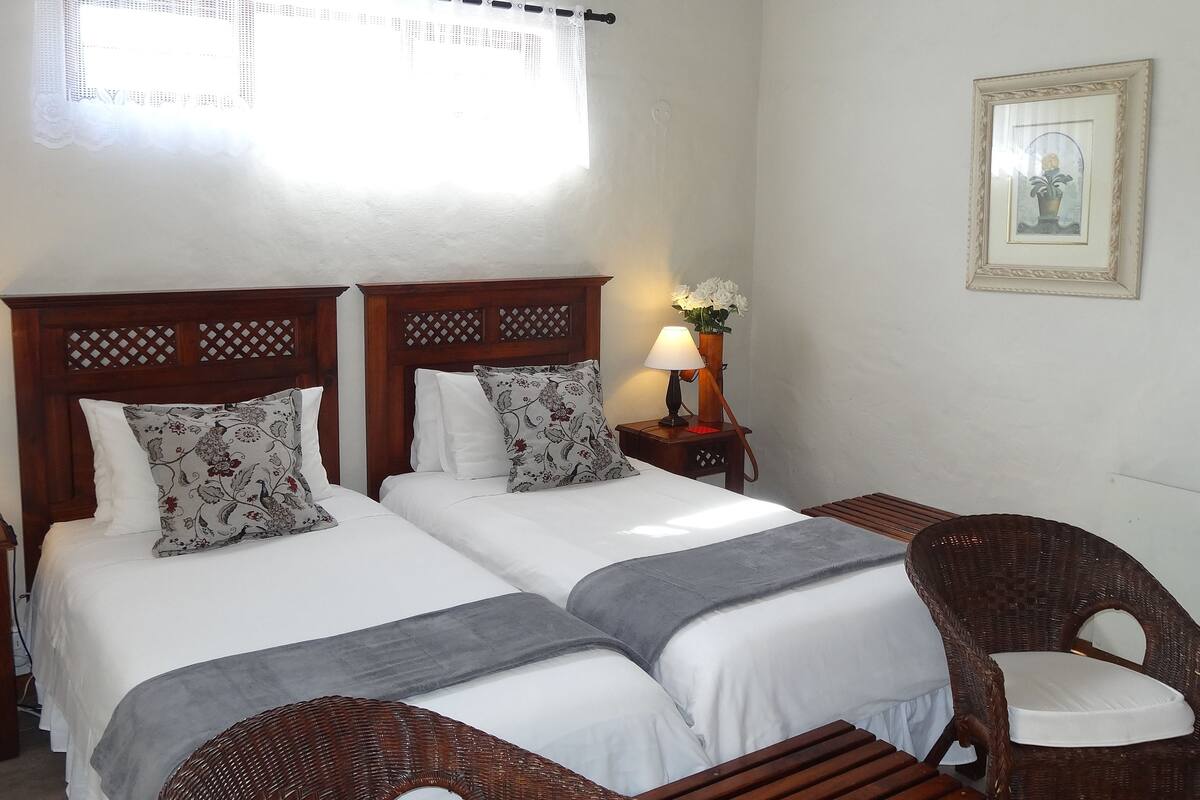 A double bed with white linens and floral-patterned pillows, a bedside table with a lamp, and a wicker chair.