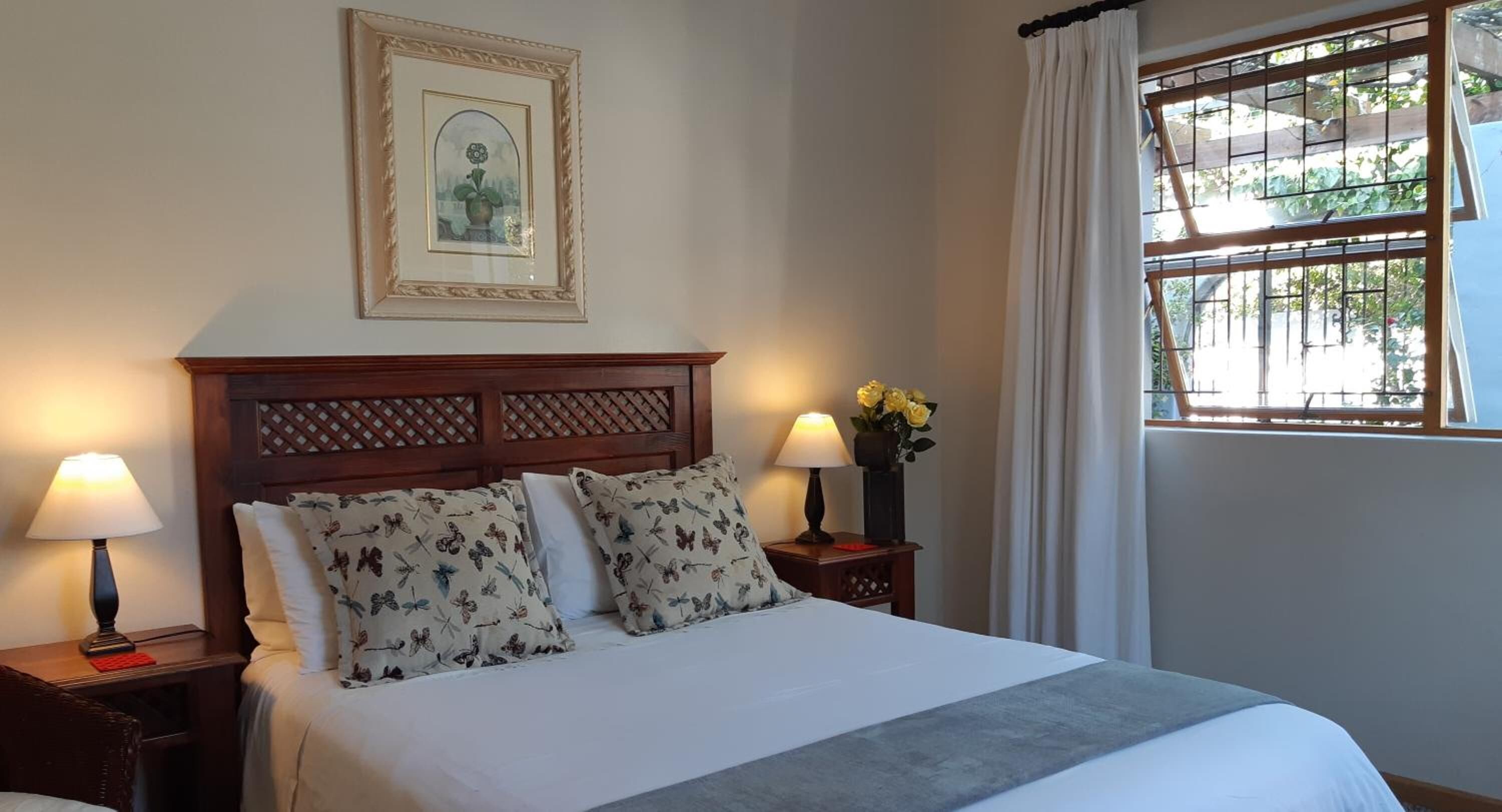 Una cama bien hecha con almohadillas florales, un cabecero de madera, un cuadro enmarcado en la pared y una ventana con cortinas.