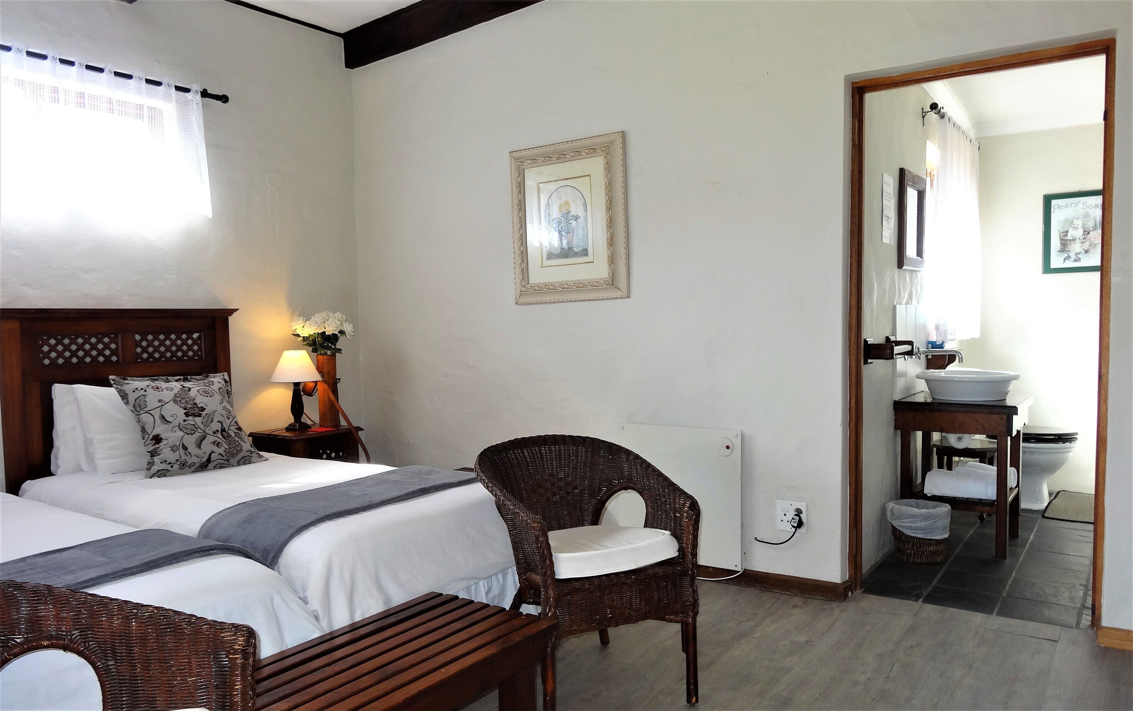 A hotel room with two beds, a wicker chair, a bedside table with a lamp, a framed picture on the wall, and a bathroom visible through an open door.