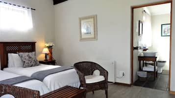 A hotel room with two beds, a wicker chair, a bedside table with a lamp, a framed picture on the wall, and a bathroom visible through an open door.