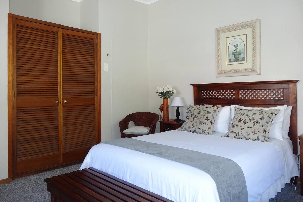 A bedroom with a large wooden wardrobe, a bed with floral pillows, a nightstand with a lamp, and a chair.