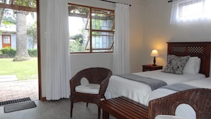 A room with a bed, two wicker chairs, a wooden bench, a nightstand with a lamp, and a window with curtains. - Be My Guest Lodge (Cape Town)