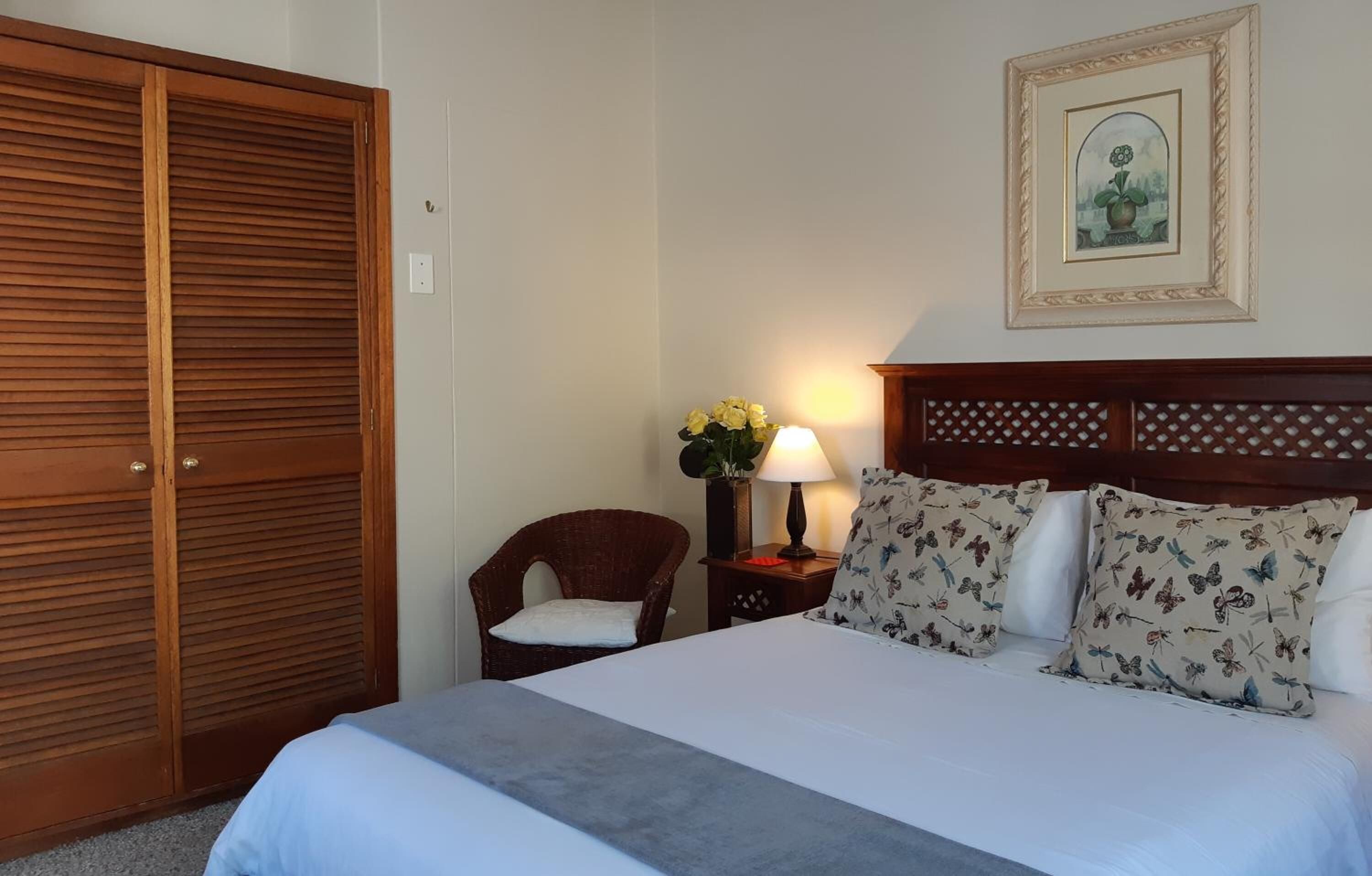 A hotel room with a bed, a wooden wardrobe, a chair, a nightstand with a lamp, and a framed picture on the wall.