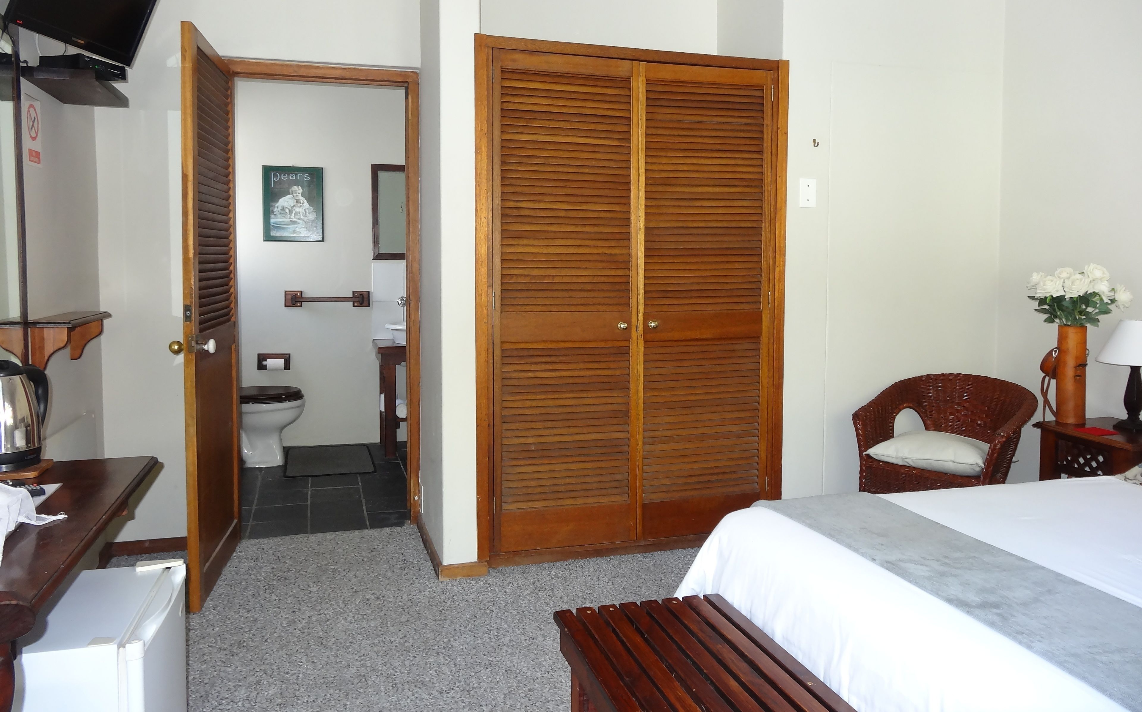 A bedroom with a wooden wardrobe, a bed, a chair, a small table with a vase of flowers, and a view into the bathroom.