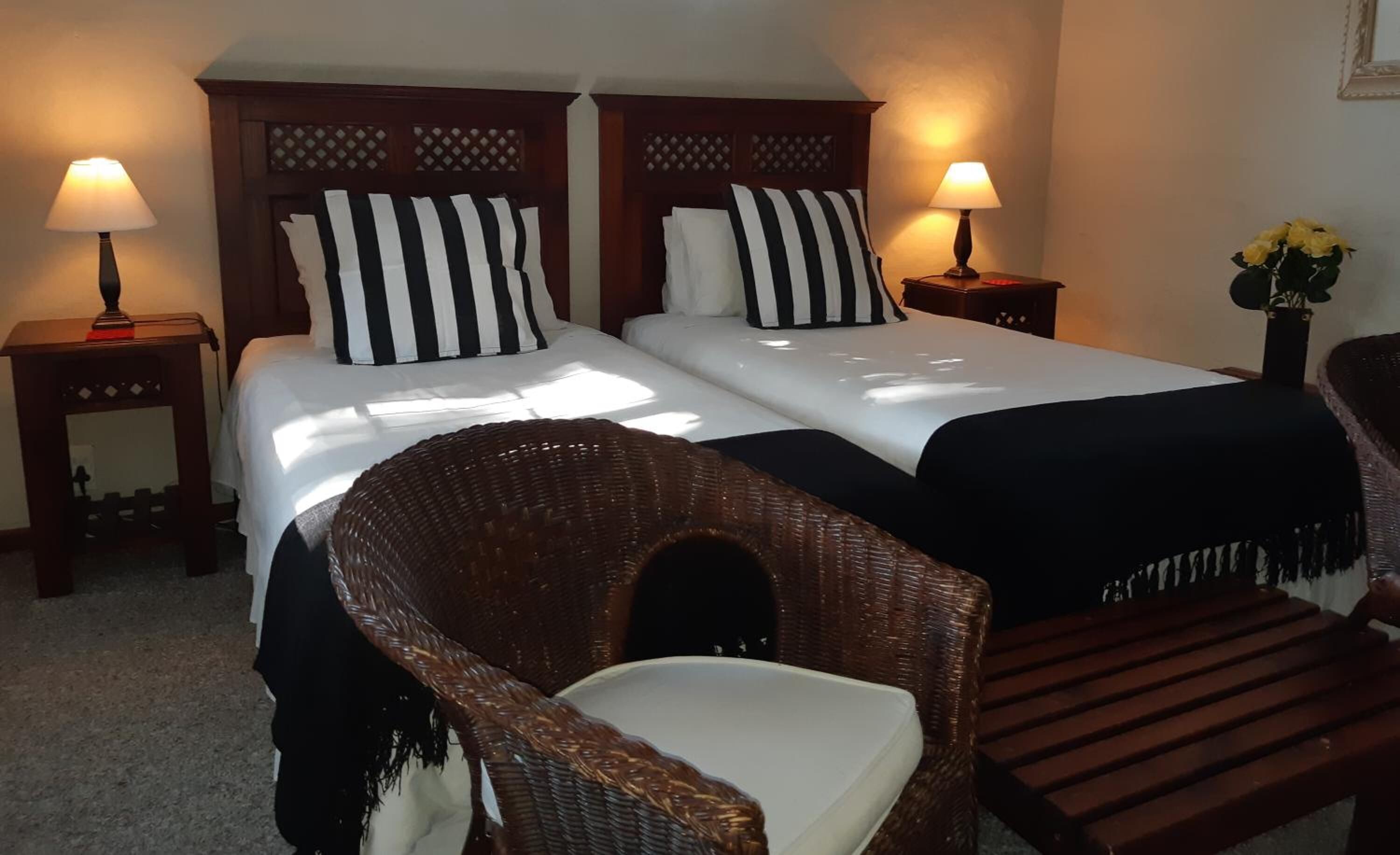 A double bed with black and white striped pillows, two bedside tables with lamps, a wicker chair, and a wooden bench.