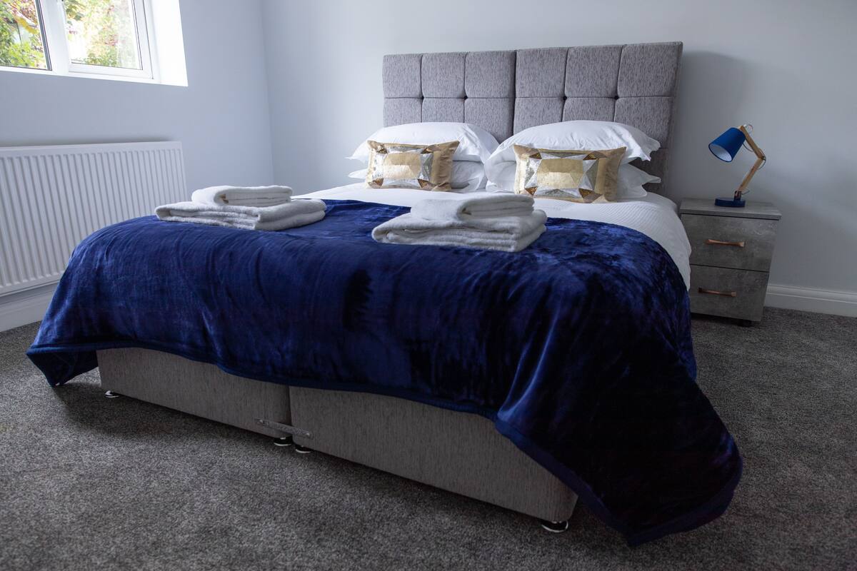Un lit bien fait, recouvert d’une couverture bleue, avec des oreillers blancs et des serviettes pliées. On y voit une tête de lit grise, une table de chevet avec une lampe et un radiateur placé sous la fenêtre.