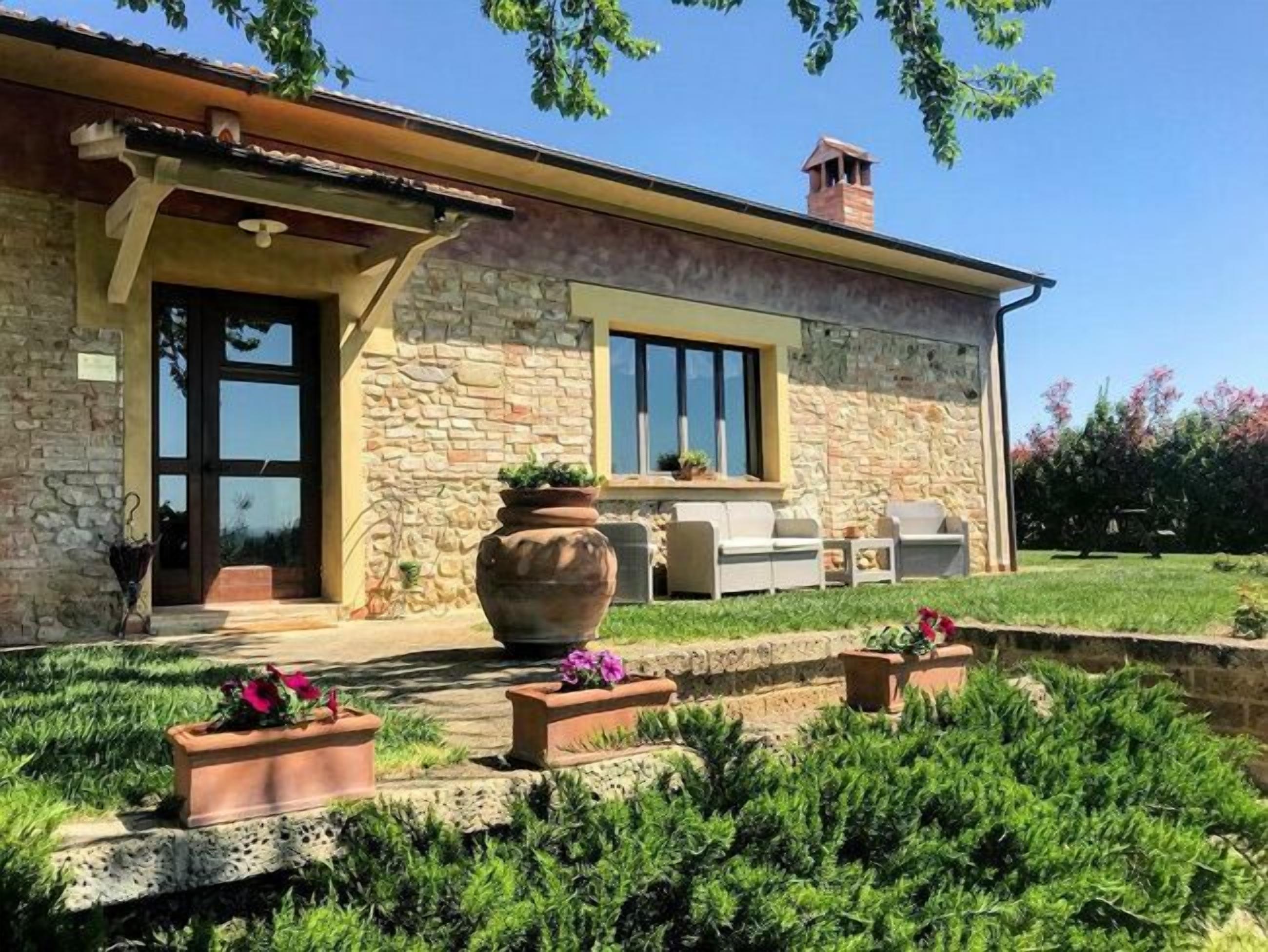 A stone house with a large window, outdoor seating, and a garden with potted plants.