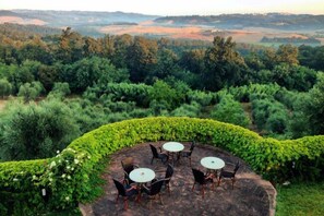 An outdoor seating area with round tables and chairs, surrounded by neatly trimmed hedges and a backdrop of rolling hills and dense trees. - Agriturismo L'Antica Fornace (Peccioli)