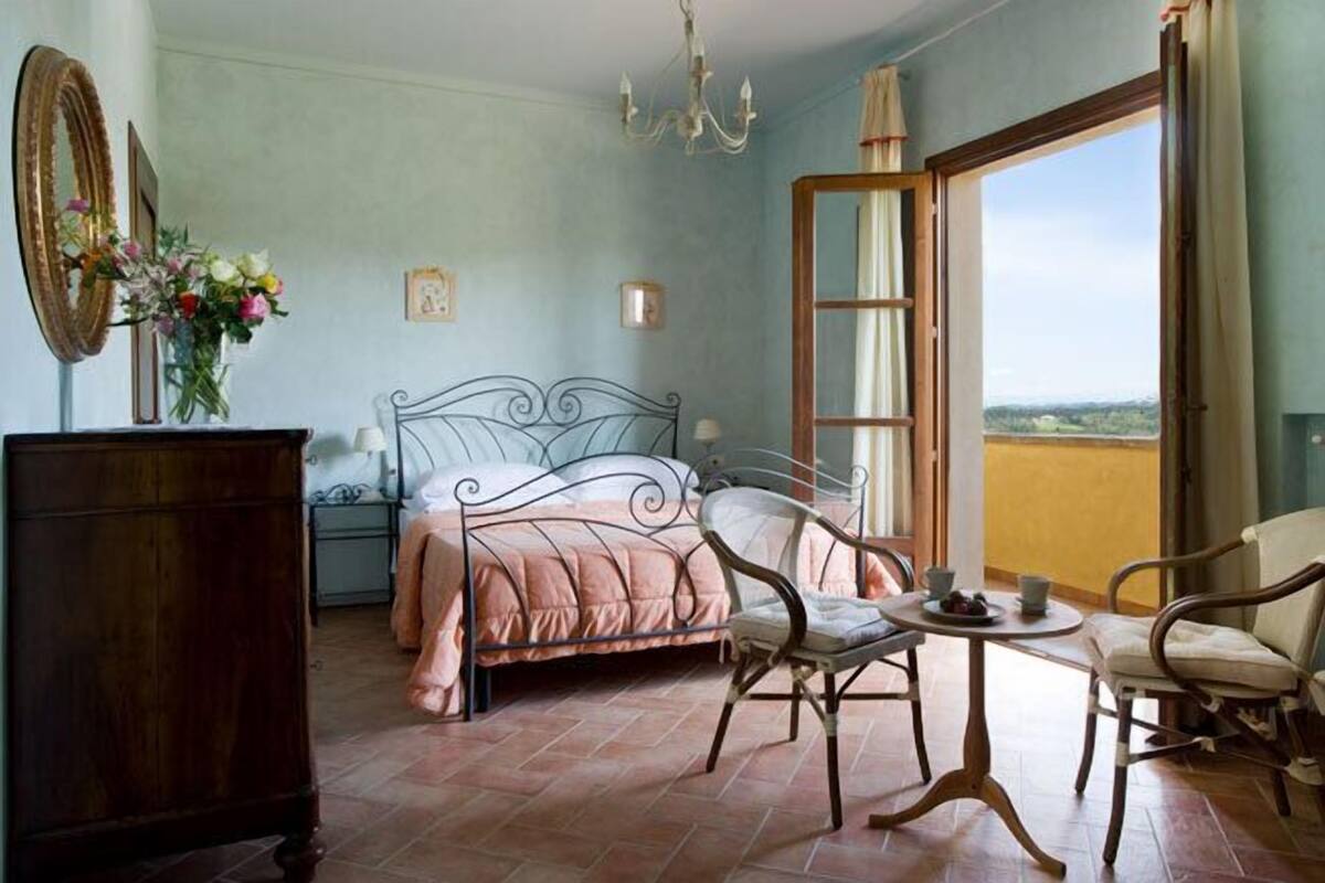 A bedroom with a bed, a chair, a table, a dresser, a mirror, and a window with a view.