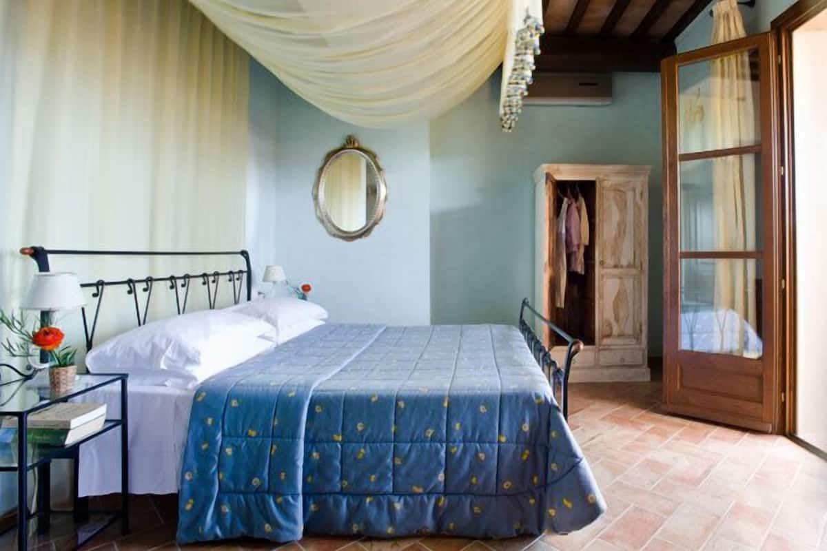A bedroom with a bed, a nightstand, a mirror, a wardrobe, and a door leading to another room.