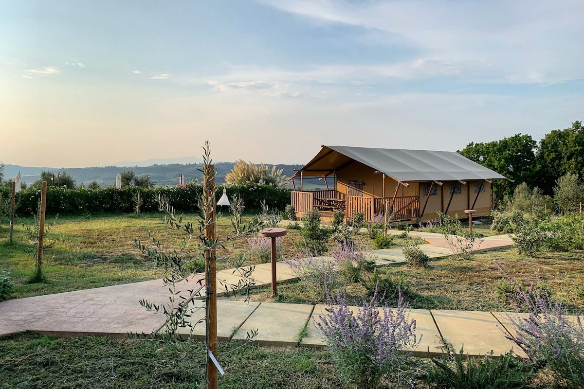 A modern, single-story wooden cabin with a large porch and a covered outdoor area, surrounded by a well-maintained garden and a scenic view of distant hills.